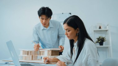 Profesyonel Asyalı erkek mimar ev modelinin uzunluğunu ölçmek için cetvel kullanırken, beyaz tenli genç meslektaşı ev modeliyle toplantı masasındaki verileri analiz etmek için dizüstü bilgisayar kullanıyor. Tertemiz..