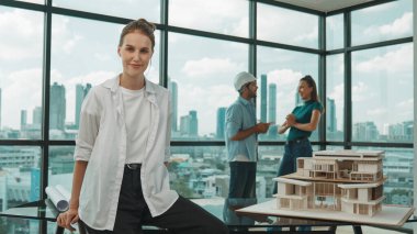 Genç ve güzel proje yöneticisi kameraya gülümserken mühendis ekibi ev tasarımı hakkında konuşuyor. Modern ofiste ev modelinin yanında otururken kameraya bakan yetenekli bir iş kadını. İzleme