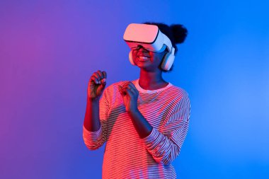 Kullanıcı deneyiminde VR ile sunum yapan genç Afrikalı bir kadın, üç boyutlu teknoloji yenilikçiliği hologram teknolojisi uygulamasındaki çevrimiçi neon stüdyo ışıklandırma arka planı aracılığıyla sunuluyor. Entrika.