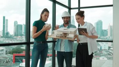 Proje yöneticisi ev modelini elinde tutarken, mimar mühendis takımı da ev tasarımı veya bina inşaatı hakkında fikir alışverişinde bulunuyor. Şehrin penceresinin yanında iç mimar duruyor. İzleme