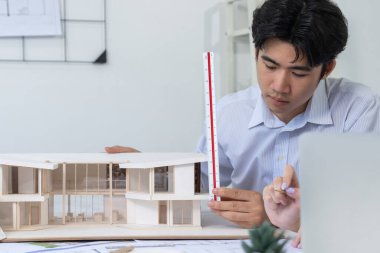 Professional male asian architect using ruler to measure house model length while young beautiful caucasian colleague using laptop to analyzed data on meeting table with house model. Immaculate. clipart