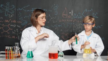 Kafkas öğretmeni, bilim laboratuarındaki renkli çözeltiden bahsetti. Uzman eğitmen, karatahta yazılı kimyasal teoride STEM bilim sınıfında deney yapan zeki çocuğu destekliyor. Etkinlik.