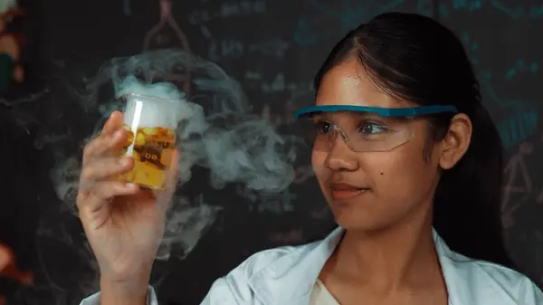 stock image Closeup of smart highschool student inspect colored solution at blackboard. Beautiful scientist holding beaker and looking at chemical liquid while standing in front of blackboard at lab. Edification.