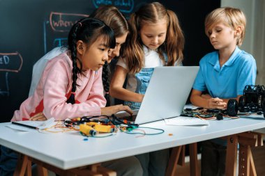 Uzaktan kumandalı araba programlamayı öğrenen yetenekli çocuklar. Pembe elbiseli liseli kız kodlama için dizüstü bilgisayar kullanıyor. Öğretmen onu izlerken diğer çocuk kodunu izliyor. Anakart ve elektrik kablosu da masada. Etkinlik.
