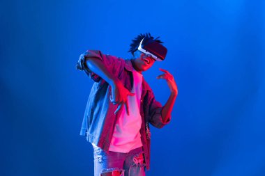 Gülümseyen Afrikalı genç Amerikalı sanal gerçekliği dijital fütüristik teknolojiye bağlayan pembe mavi neon duvarda hologram oyunlarıyla VR dansına bakıyor. Entrika.