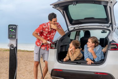 Aile gezisi plajda elektrikli arabayla seyahat ediyor, güzel bir aile bagajda oturuyor, EV araba aküsünü yeşil ve sürdürülebilir enerjiyle şarj ediyor. Aile seyahati ve çevre dostu araba. Kalıcı