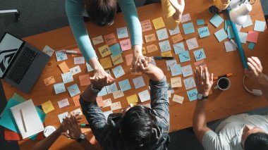 Top view of business team giving high five to celebrate project while standing at table with sticky notes and laptop. Aerial view of designer clapping hands to congratulate successful idea. Symposium.