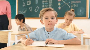 Çekici beyaz çocuk okulda matematik okurken kameraya bakıyor. Çeşitli şirin öğrenciler test yaparken öğretmen Asyalı çocuklara soru sorup tahtaya kağıt doğrultuyor. Pedagoji.