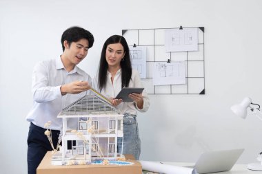 Güzel, genç, beyaz bir proje yöneticisi Asyalı mühendise, mühendislik masasında ev modelini ölçerken tablet kullanarak bina detaylarını anlattı. Tertemiz.