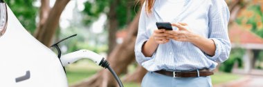 Panorama 'lı genç bir kadın EV şarj istasyonundan EV elektrikli araç aküsünü şarj ediyor ve şehir parkındaki elektriği ödemek için akıllı telefon internet bankacılığını kullanıyor. EV arabalı çevre dostu bir araç. Yükselt