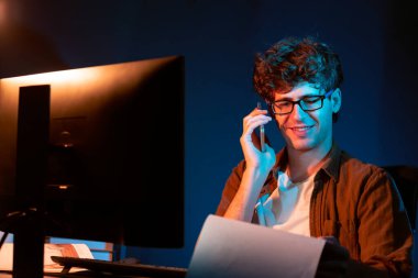 Genç iş adamı akıllı telefondan müşteriyi ya da iş arkadaşını arıyor, neon modern ofisteki yaratıcı sunum surround çalışma masasının bilgisayar ekranına bakıyor. Yeni proje kavramı. Toplayıcı.