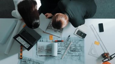 Planlara bakan, proje planı ve güvenlik kaskıyla toplantı masasında oturan mühendis ile konuşan yetenekli yöneticinin üst görüşü. Mimarların hava görüntüsü belgeyi tutar ve yürür. Hizalama.