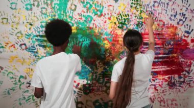 Genç ve çeşitli çocukların arka görünüşleri renkli duvarları elle boyarlar. Çekici bir lise öğrencisi renk paletini tutarken duvarı el iziyle boyuyor. Yaratıcı aktivite. Öğretim