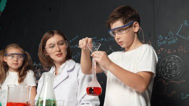 Öğretmen tavsiye verirken beyaz çocuk kimyasal sıvıyı karıştırıyor. Profesyonel eğitmen labaratuar kıyafeti giymiş, renkli solüsyonla dolu deney kabıyla sofrada çeşitli öğrencileri arıyor. Etkinlik.