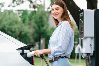 Açık yeşil şehir parkı manzaralı EV şarj istasyonundan genç kadın EV elektrikli araçları şarj ediyor. Çevre dostu şehir ulaşımı ve çevre dostu EV araba seyahati. Yükselt