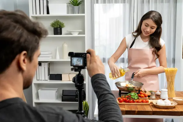 stock image Cameraman recording to woman in chef influencer host cooking spaghetti with meat topped tomato sauce surrounded ingredients recipe, presenting special dish healthy food at modern studio. Postulate.