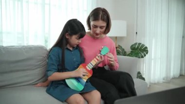 Kafkasyalı bir anne evde akustik müzik öğretirken ukulele çalan mutlu kız. Sevimli çocuk enstrüman öğreniyor. Kafkasyalı mutlu anne ve kız birlikte vakit geçiriyorlar. Pedagoji.