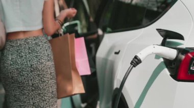 Yaz mevsiminde yeşil sürdürülebilir şehir bahçesinde ağır çekimde çalışan EV elektrikli araba yeşil sürdürülebilir şehirde yeniden şarj edilebilir elektrikli araç iç organları için alışveriş yapan genç çiftlerin yaşam tarzını gösteriyor.