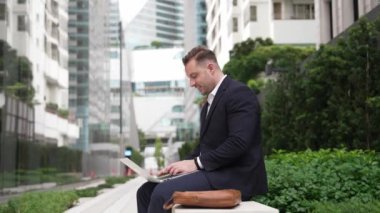 Yeşil Şehir 'deki akıllı proje yöneticisine rapor yazmak için dizüstü bilgisayarı üzerinde çalışıyor. İş adamı analiz verilerine ve beyin fırtınası fikrine bankta otururken bilgisayar kullanarak bakıyor. Urbane.