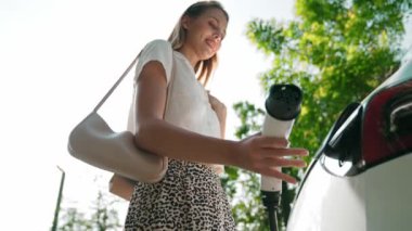 Yavaş çekim güneş ışığı arka plan aydınlatması EV elektrikli arabasıyla gezen kadın yazın yeşil sürdürülebilir şehir bahçesinde şarj oluyor. Elektrikli araç iç organlarından yeşil kentsel sürdürülebilirlik yaşam tarzı