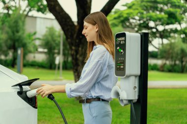 Açık yeşil şehir parkı manzaralı EV şarj istasyonundan genç kadın EV elektrikli araçları şarj ediyor. Çevre dostu şehir ulaşımı ve çevre dostu EV araba seyahati. Yükselt