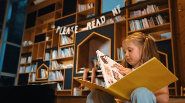 Genç, zeki, beyaz bir kız kütüphanede otururken kitap okuyor. Zeki çocuk öğrenme, okuma, kütüphanede kitap açma. Etkileyici çocuk geçmişi bulanık sayfayı çeviriyor. Etkinlik.