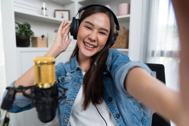 Sunucu kanalı genç ve güzel Asyalı kız akıllı telefonuyla selfie çekiyor. Modern stüdyo ofis konseptinde kulaklık takıyor. Sahne arkası yayıncısı sosyal medyada canlı yayın yapıyor. Taktik.