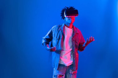 Gülümseyen Afrikalı genç Amerikalı sanal gerçekliği dijital fütüristik teknolojiye bağlayan pembe mavi neon duvarda hologram oyunlarıyla VR dansına bakıyor. Entrika.