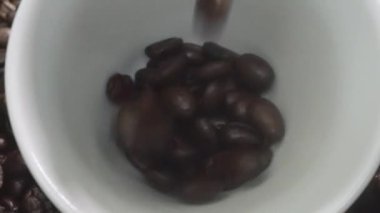 Kahve fincanının içine kahve çekirdeği yığını ile çevrili bir şekilde düşüyor. Beyaz fincana aromatik fasulye döküyorum. Kahvenin üst görüntüsü siyah arka planlı cama düşer. Geliştirilebilir.