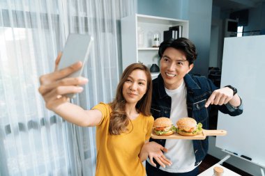 Gülümseyen sunucu şefler akıllı telefonla selfie çekerek sosyal medya kanalından canlı yayında yemek programında çift büyük hamburger yapıyorlar. İnfobahn.