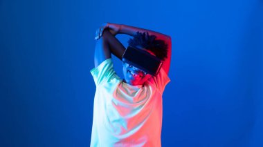 Genç Afro-Amerikalı sporcu VR 'ı giyip kollarını üst tarafa uzatarak mavi neon ışıklı duvara poz vererek metaevren dünyasındaki dijital fütüristik teknolojiyi sanal gerçekliğe bağlıyor. Entrika.