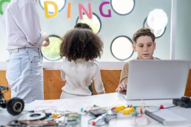 Sınıftaki çocuklar BTMM sınıfında mühendislik öğreniyor. Okul çocuğu sandalyede oturuyor ve uzaktan kumandalı arabayı dizüstü bilgisayarla kodluyor. Öğretmen ve diğer çocuklar etrafında. Not defteri, elektrik kablosu. Etkinlik.