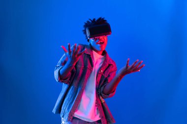 Gülümseyen Afrikalı genç Amerikalı sanal gerçekliği dijital fütüristik teknolojiye bağlayan pembe mavi neon duvarda hologram oyunlarıyla VR dansına bakıyor. Entrika.