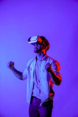 Profesyonel oyuncu, renkli neon ışıklı arka planda VR gözlük takarken oyunu kazanmasını kutlamak için elini havaya kaldırdı. Beyaz adam teknolojik yenilikleri kullanıyor ve giriyor. Sapma.