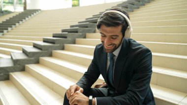 Gülümseyen iş adamı ortağını beklerken akıllı saate bakıyor. Modern alışveriş merkezinin merdivenlerinde otururken proje yöneticisi müzik dinliyor. Alışveriş merkezinde takım elbiseli mutlu bir adam bekliyor. Sevinçli..