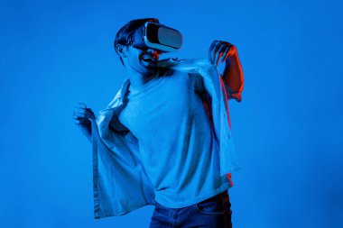 Yakışıklı, dans eden ve müziğe hareket eden bir adam. VR gözlüğü ve neon ışıklı arka plan kullanarak. Beyaz erkek oyuncu, teknolojik yenilikler giyerek pop müzik dinlerken ayakta duruyor. Sapma.