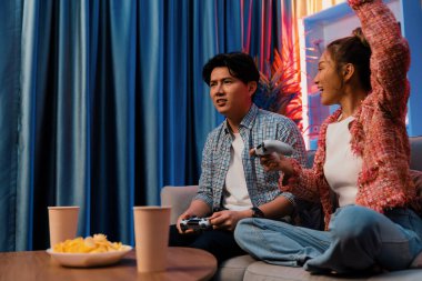 Mutlu çiftin oturduğu kanepe joysticklerle video oyunu oynuyor, meydan okuyan gülümseyen kadın, evdeki modern neon ışıklı oturma odasındaki mücadeleyi kazanmak için yumruk kaldırıyor. İnfobahn.