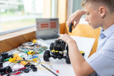 Sarı saçlı okul çocuğu, BTMM sınıfında robotik araç tamir eden bir tornavida kullanıyor. Masaya dizüstü bilgisayar, kumanda, elektrik kablosu, şarj aleti ve robot araç koyun. Öğretim.