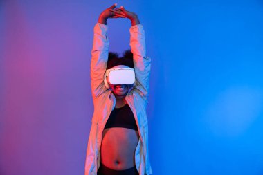 VR giyen genç Afrikalı spor kadın, dijital fütüristik teknolojiyi sanal gerçekliğe bağlayan parlak mavi neon ışıkları üzerinde poz veriyor. Entrika.