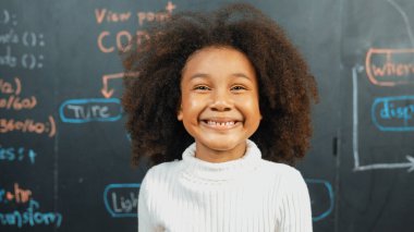 Genç Afrikalı bir kızın karatahtaya bakarken kameraya bakışı. BTMM teknoloji sınıfında arkada yazılı kamera sistemi koduna gülümseyen bir öğrenci. Etkinlik.