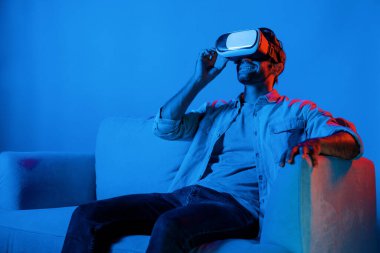 Adam film izlemek ve kanepede neon ışıkla oturmak için metaevrene girmek için VR camına bakıyor. Kafkasyalı ergen günlük giysiler giyiyor ve sanal gerçeklik simülasyonu programı takıyor. Sapma.