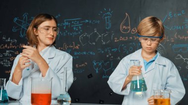 Kafkas öğretmeni, bilim laboratuarındaki renkli çözeltiden bahsetti. Uzman eğitmen, karatahta yazılı kimyasal teoride STEM bilim sınıfında deney yapan zeki çocuğu destekliyor. Etkinlik.