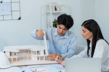 Profesyonel Asyalı erkek mimar ev modelinin uzunluğunu ölçmek için cetvel kullanırken, beyaz tenli genç meslektaşı ev modeliyle toplantı masasındaki verileri analiz etmek için dizüstü bilgisayar kullanıyor. Tertemiz..