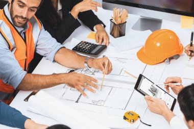 Mimari proje üzerinde birlikte çalışan çeşitli inşaat mühendisi ve müvekkil grupları, inşaat planını gözden geçirip toplantı masasında plan oluşturuyorlar. Tedbirli
