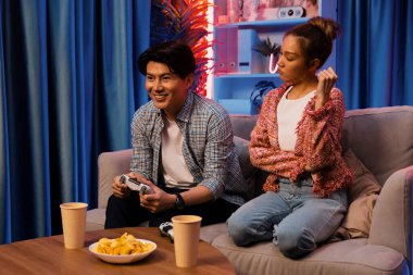 Mutlu çiftin oturduğu kanepe joysticklerle video oyunu oynuyor, meydan okuyan gülümseyen adam, evdeki modern neon ışıklı oturma odasında kazanmak için yumruğunu kaldırıyor. İnfobahn.