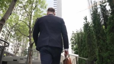 Yeşil şehirde bavulunu tutarken resmi takım elbiseli beyaz bir müdürün merdivenlerden çıkışının arka görüntüsü. Profesyonel bir iş adamı iş yerine giderken aynı zamanda Eko Şehri 'nde merdivenleri çıkıyor. Urbane..