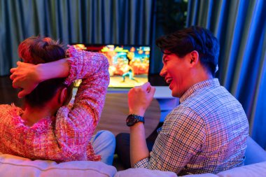 joystickli bir çift, TV ekranında dövüş oyunu oynuyor, erkek, kız arkadaşıyla yarışma seviyesini geri kazanıyor, modern evdeki neon ışıklı oturma odasında kanepeye oturuyor. İnfobahn.