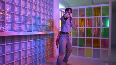 Gece kulübünde kameraya bakarken güneş gözlüklü Asyalı dansçı serbest dans ediyor. Mutlu sokak dansçısı dans ediyor ve renkli parlak neon ışığı ya da yol gösteren ışıkla ayak seslerini gösteriyor. Mutluluk.