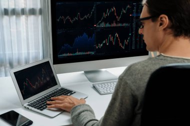 Stresli iş adamı günü modern ofisteki dinamik borsa grafiğine bakıyor, veri tabanı bilgisayarını ve bilgisayarını evden çalışırken karşılaştırıyor. Bireysel.