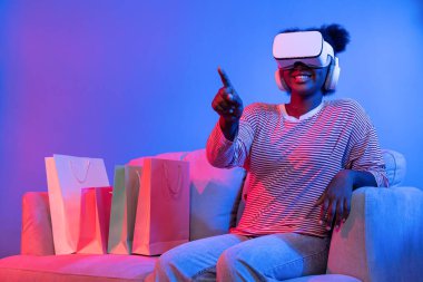 VR 'lı genç Afrikalıların alışverişinin tadını çıkarın. İnternette hologram uygulaması, güzellik ürünleri, neon ışıklandırma, oturma odasındaki koltukta alışveriş torbası bulunan canlı dijital fütüristik teknoloji. Entrika.